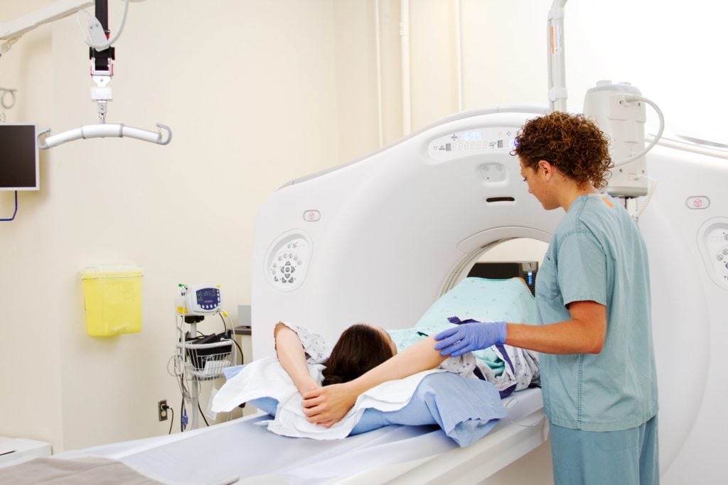 A healthcare professional prepares a patient for a CT scan. The patient lies on their back with arms above their head, sliding into the large, circular CT scanner in a medical examination room.