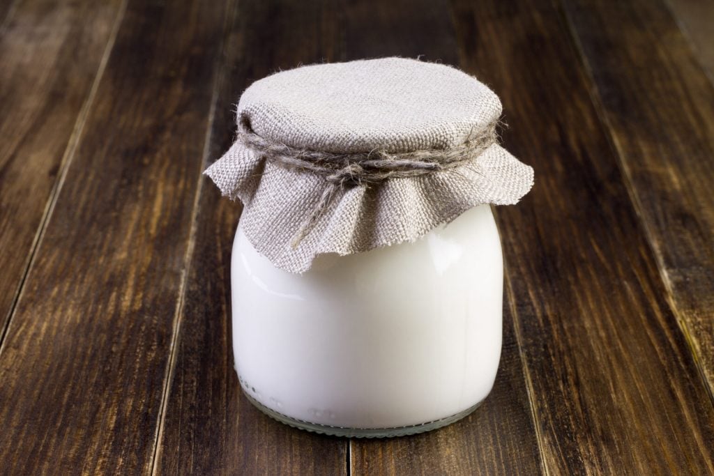 A glass jar filled with white liquid, covered with a piece of beige fabric tied with twine, sits on a wooden surface.