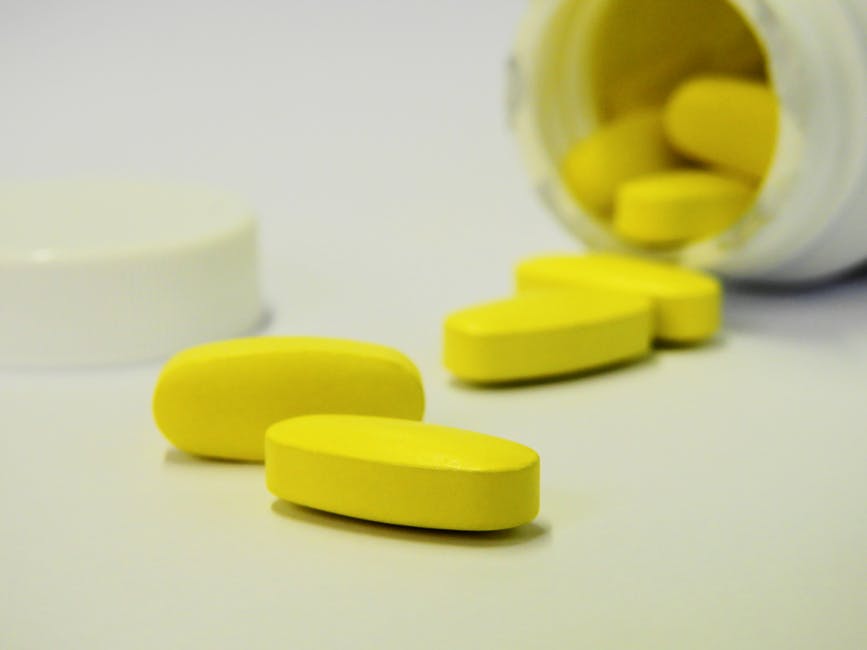 Five yellow tablets are scattered on a white surface, with some spilling out from an open white plastic bottle lying on its side. The bottle cap is placed nearby.