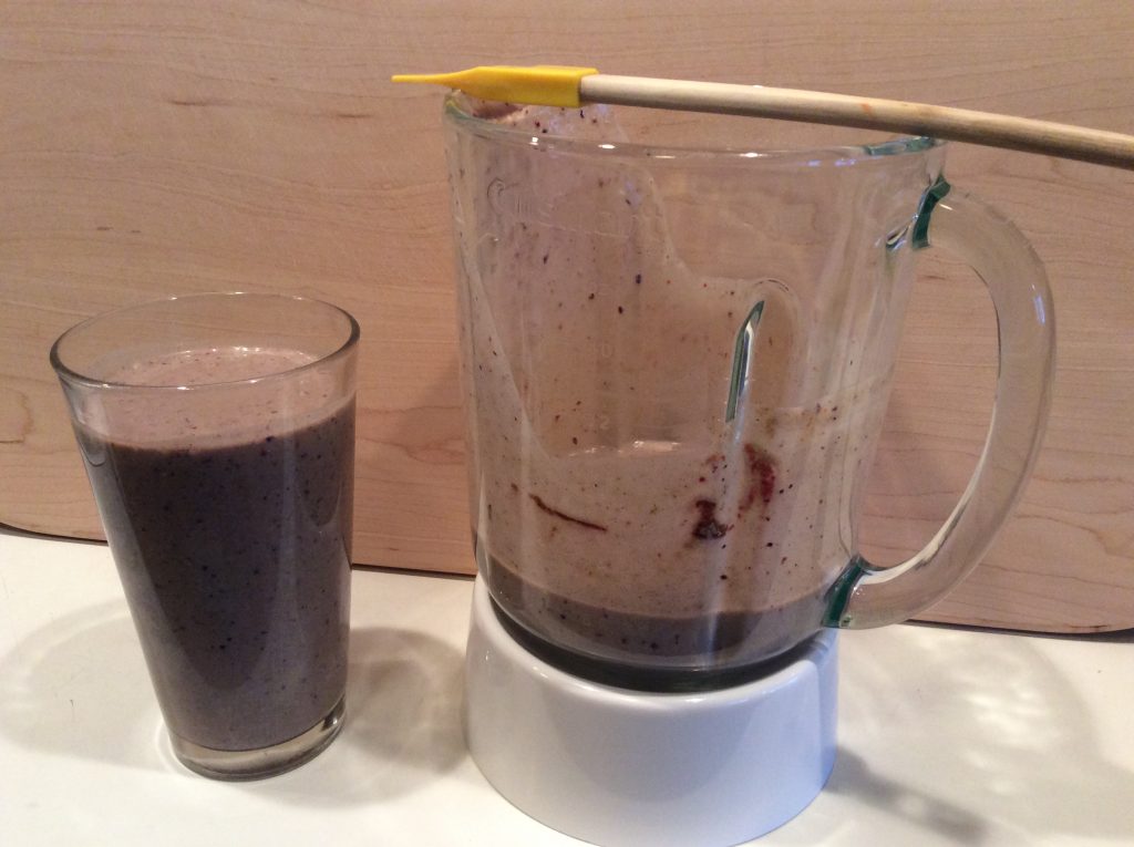 A glass of purple anticancer smoothie sits next to a blender containing leftover smoothie. A spatula rests across the top of the blender. The background is light wood, highlighting this nutritious smoothie recipe.