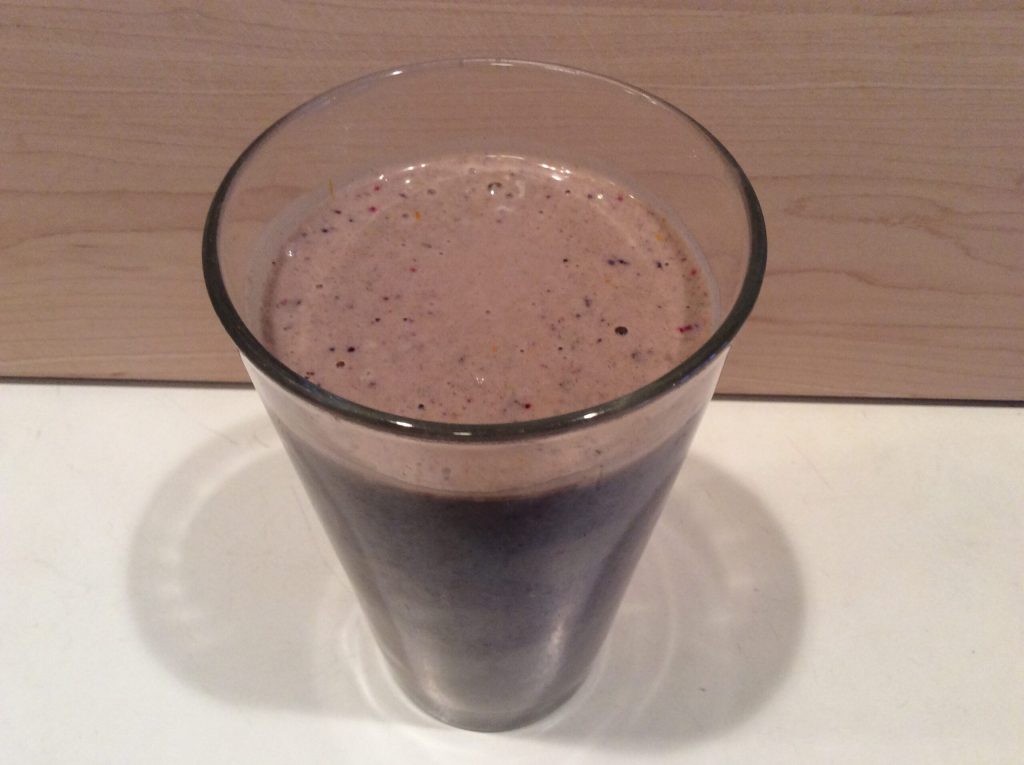 A clear glass filled with a thick, purple-brown anticancer smoothie containing visible specks of fruit, placed on a white surface with a light wooden background.
