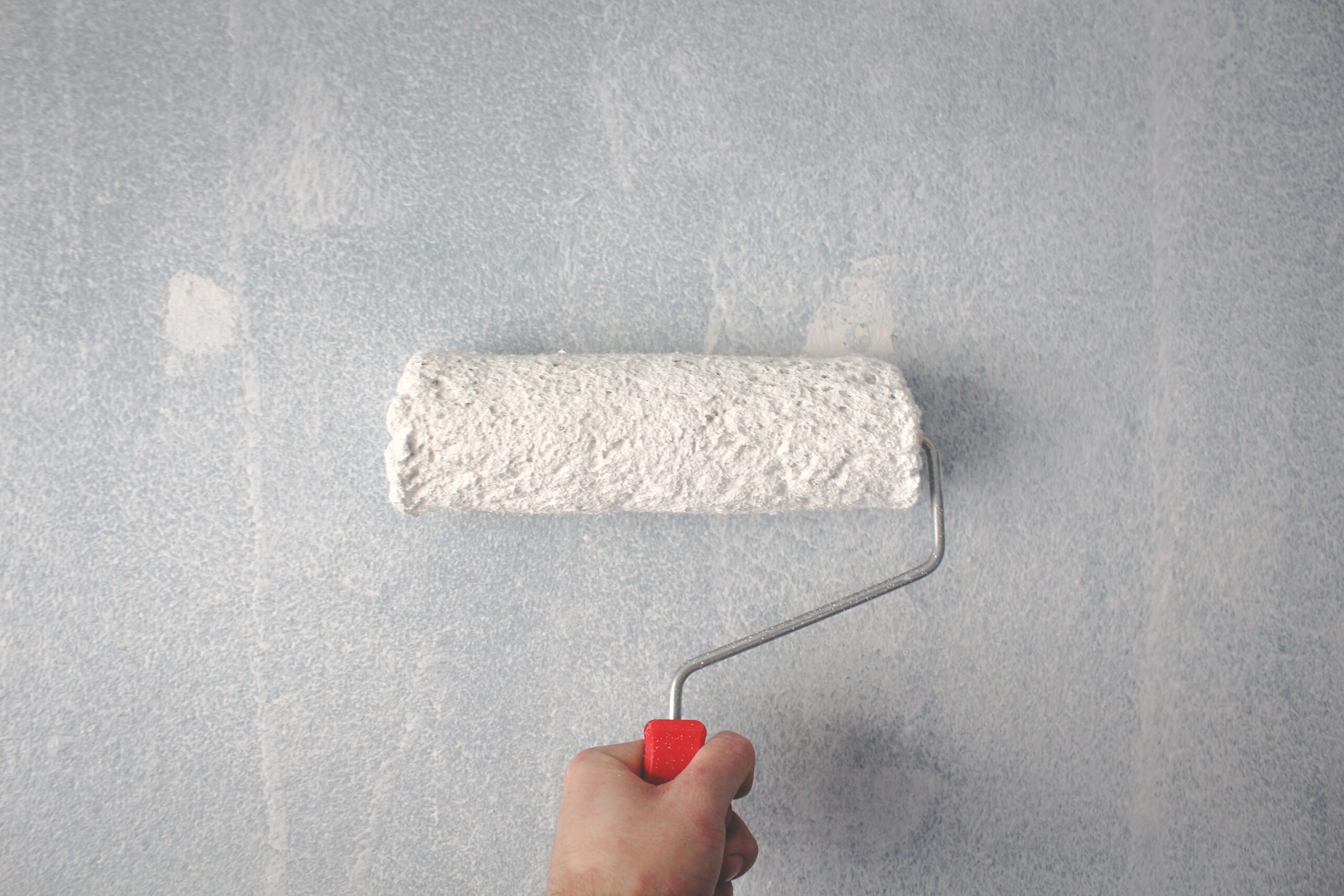 A hand holding a paint roller with white paint, applying it to a light gray wall. The partially painted surface highlights the importance of non-toxic painting for improved paint safety during home projects.