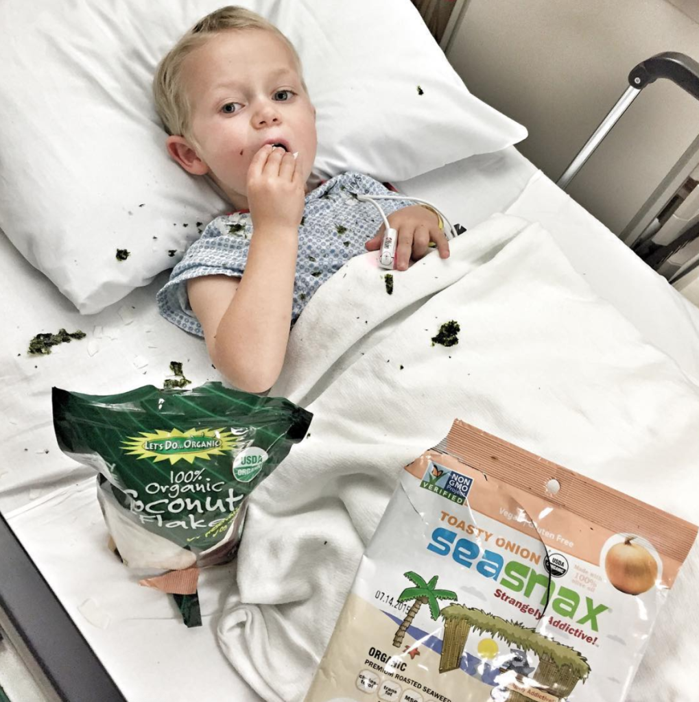 A young child lies in a hospital bed, eating snacks to help minimize side effects. Two open bags, one of organic coconut flakes and the other of SeaSnax roasted seaweed, sit on the bed with pieces scattered around.