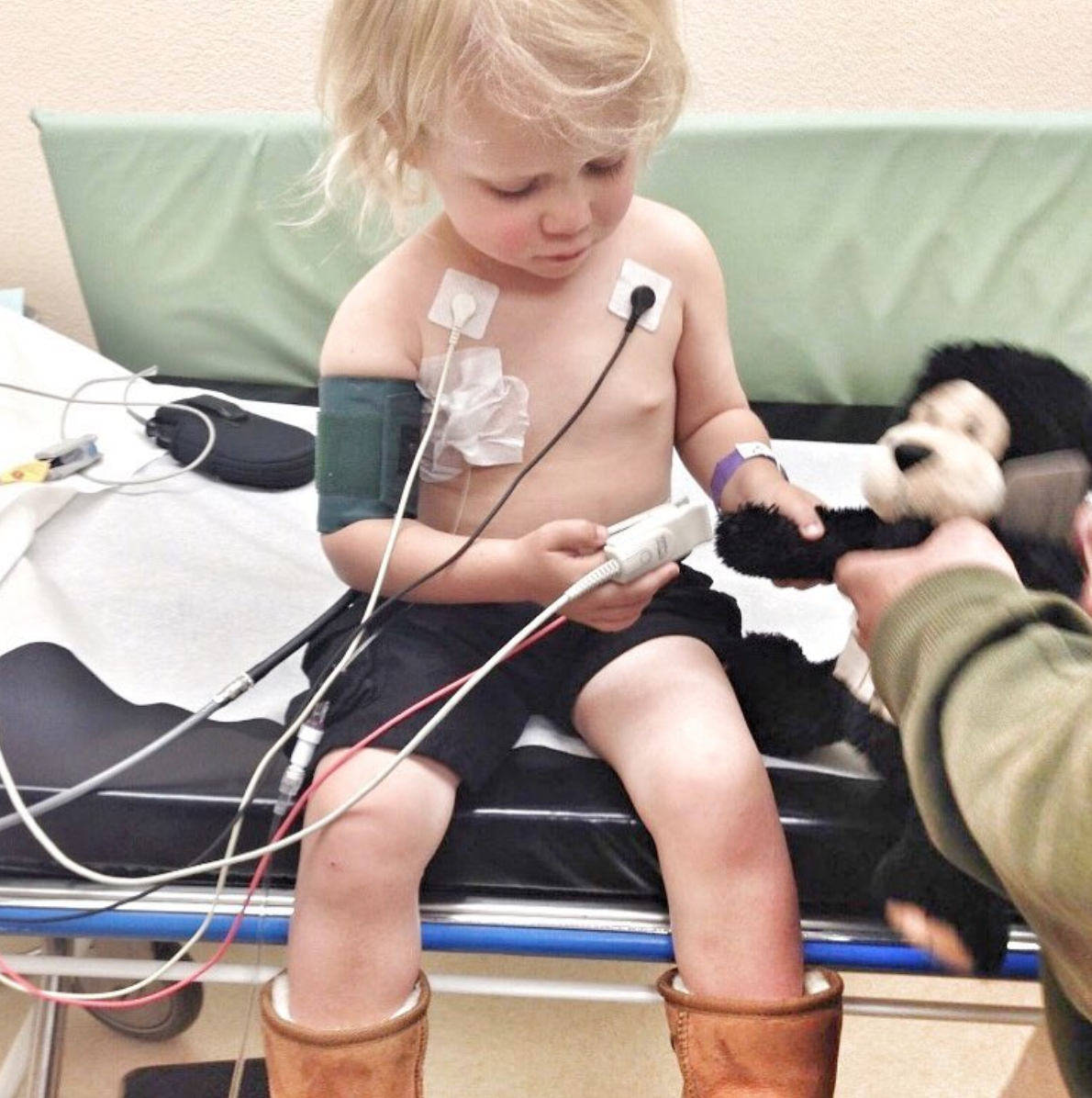 A young child sits on a hospital bed wearing medical sensors and a blood pressure cuff, holding a monitor. An adult hands the child a stuffed animal to help provide comfort and minimize side effects during treatment.
