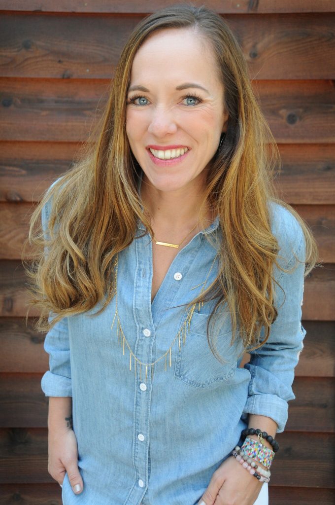 A woman with long, light brown hair smiles at the camera. She is wearing a light denim shirt, layered necklaces, and colorful bracelets, standing in front of a wooden wall after chemotherapy and surgery.
