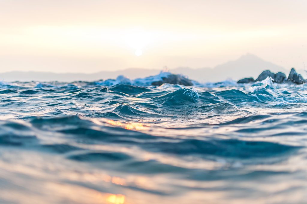 Close-up view of ocean waves at sunset with gentle ripples, reflecting soft sunlight. In the background, the sun is setting near distant mountains under a hazy sky, evoking Gerald Pollack’s concept of the fourth phase of water.