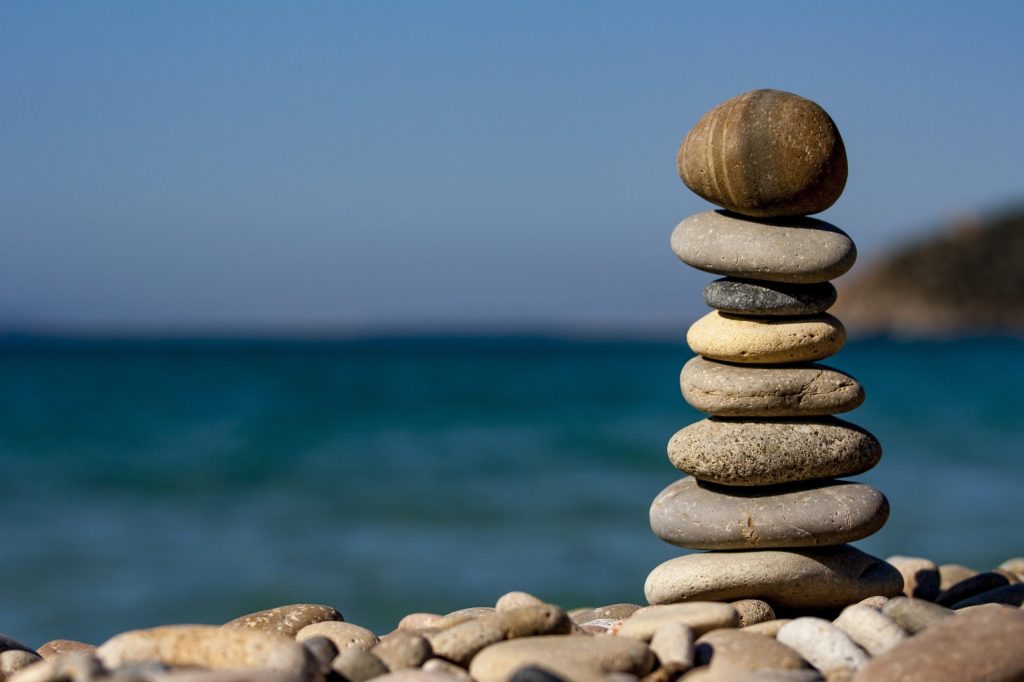 A stack of smooth, round stones balanced on a pebbly beach highlights the tranquil coastal environment, with a calm blue sea and clear sky in the background.