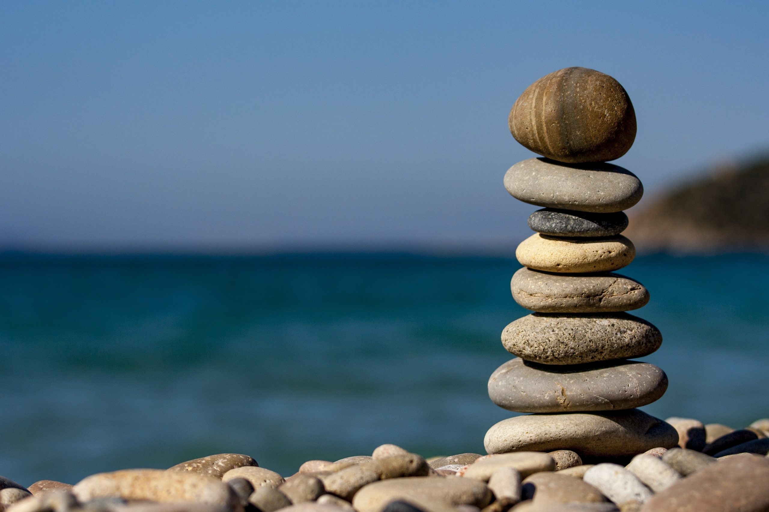A stack of smooth, round stones balanced on a pebbly beach highlights the tranquil coastal environment, with a calm blue sea and clear sky in the background.