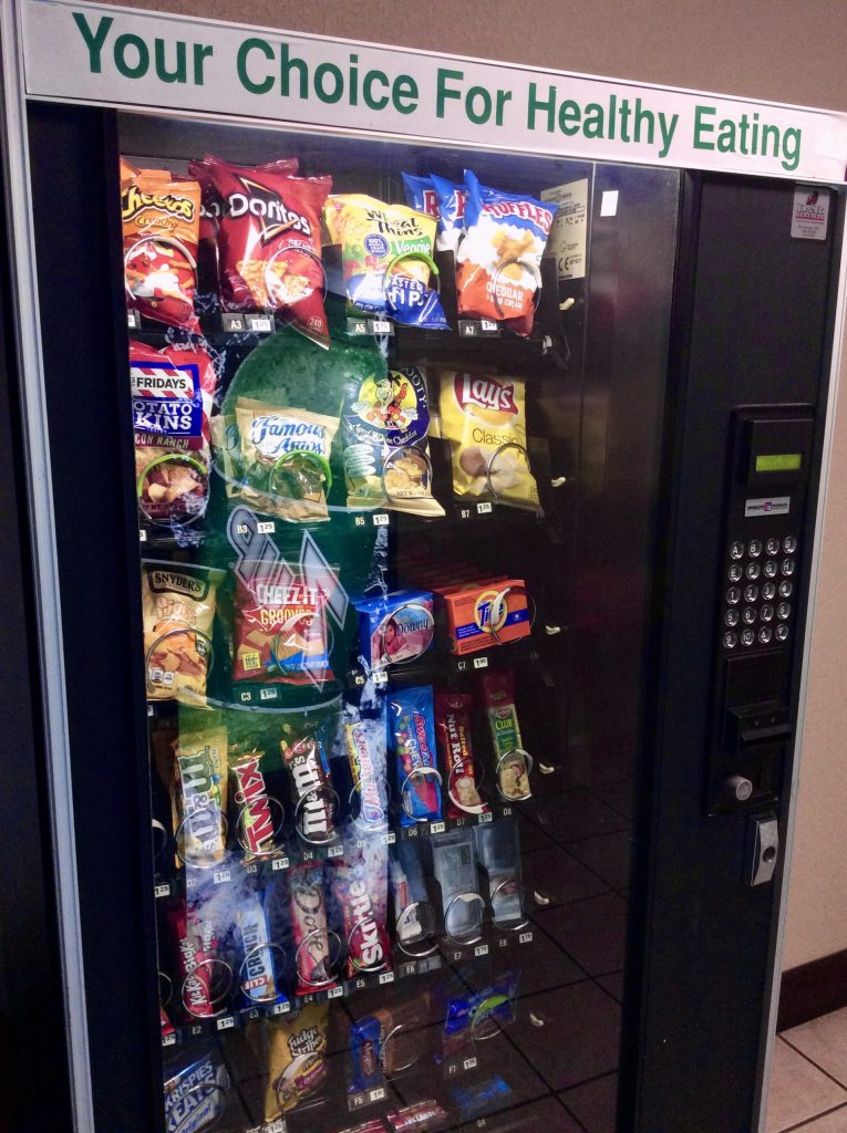 A vending machine labeled Your Choice For Healthy Eating, yet filled with chips, cookies, candy, and other fake food snacks—clearly contradicting the message of food authenticity.