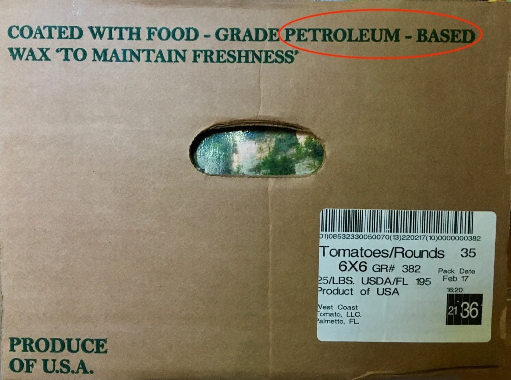 A cardboard produce box labeled COATED WITH FOOD-GRADE PETROLEUM-BASED WAX TO MAINTAIN FRESHNESS, raising questions about food authenticity, with a shipping label for tomatoes and a cutout handle showing green inside.