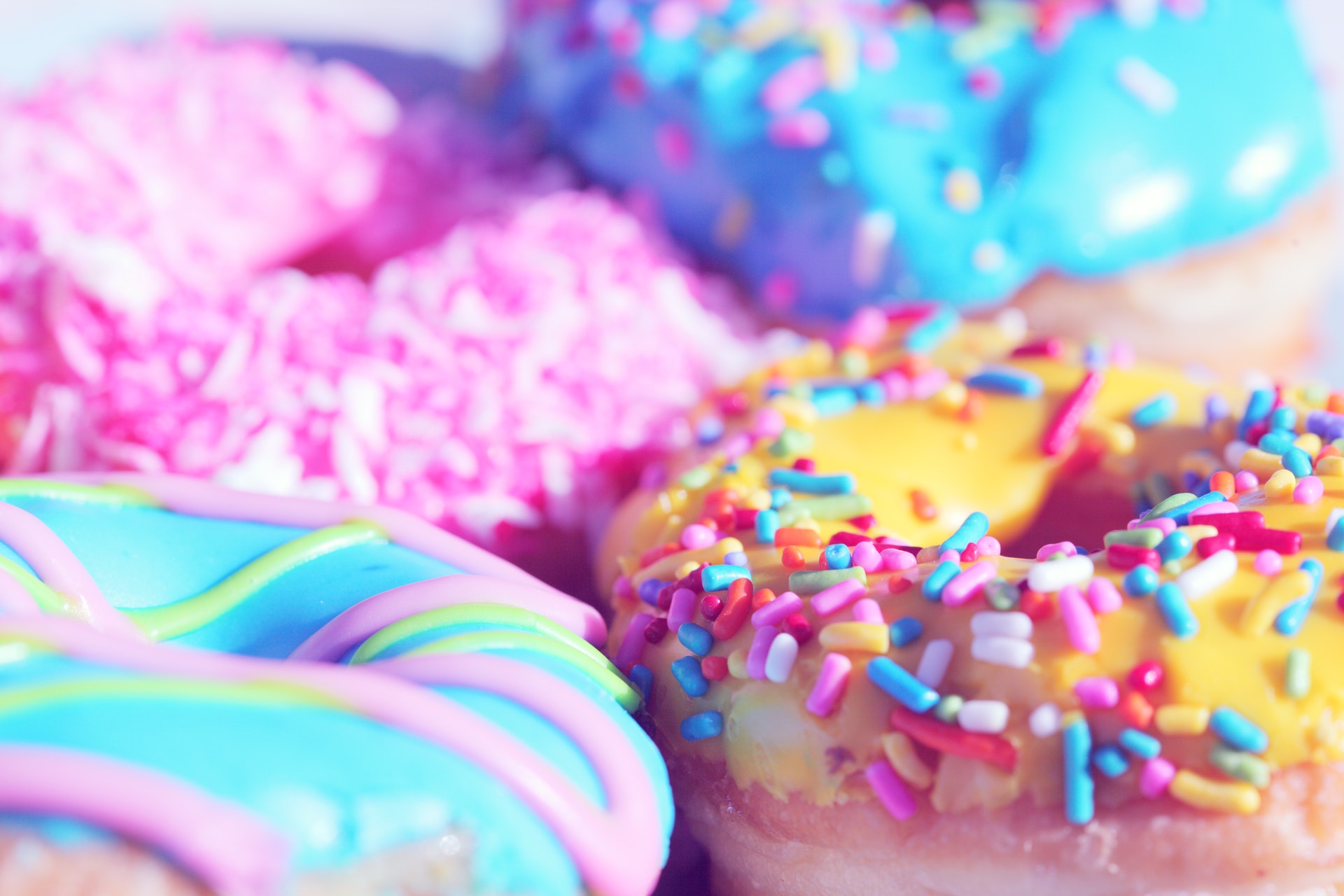 Four colorful donuts topped with bright icing and rainbow sprinkles, arranged closely together. These realistic treats showcase food authenticity, featuring pink with coconut, blue with sprinkles, yellow with sprinkles, and blue with drizzle.