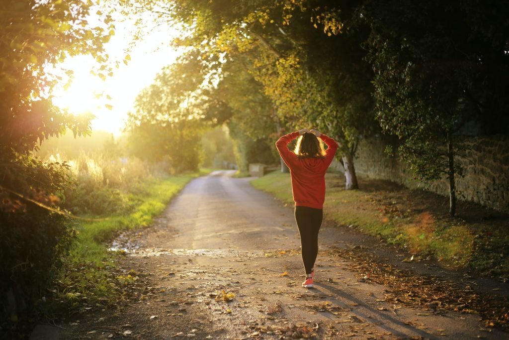 A person in a red sweatshirt walks down a quiet, tree-lined path at sunrise or sunset, with sunlight streaming through the leaves and autumn foliage scattered on the ground, generating health with each peaceful step.