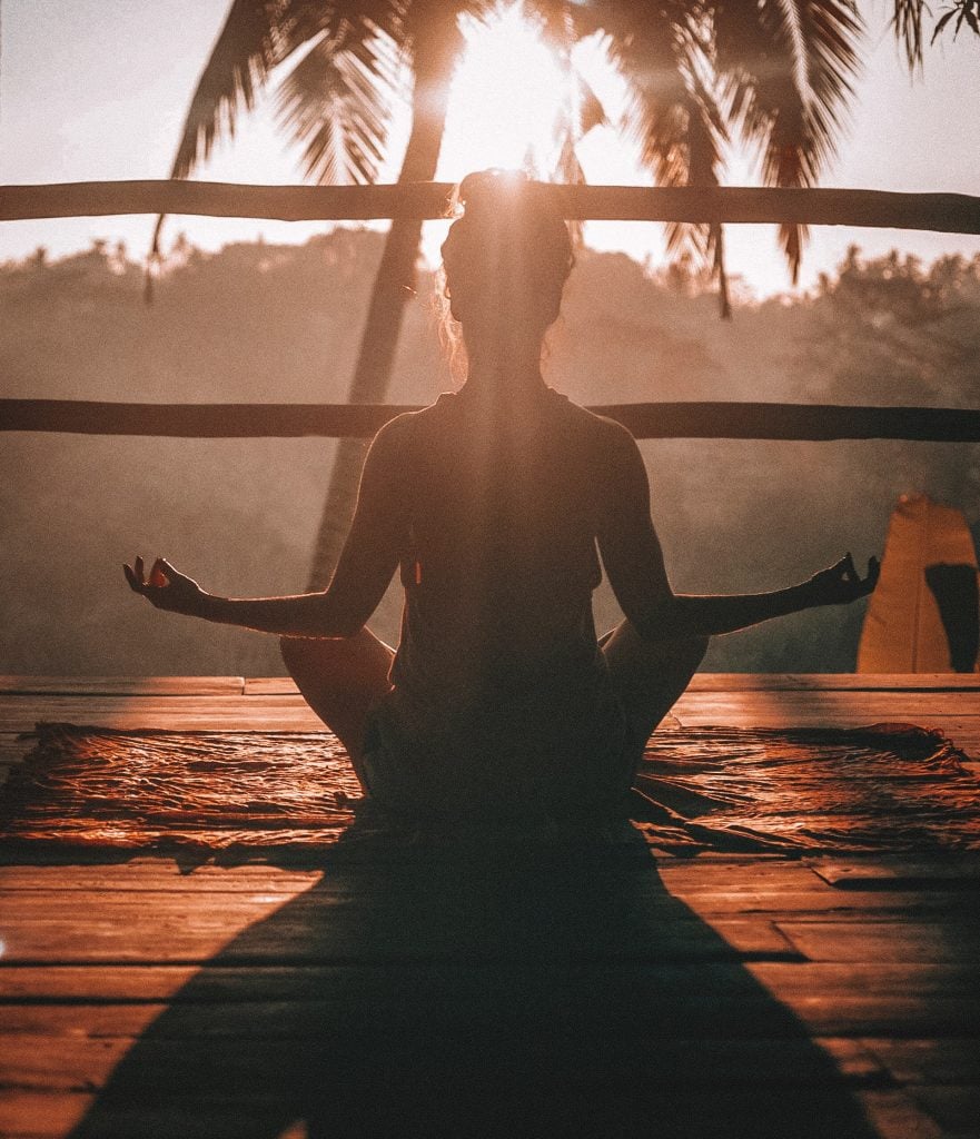 A person sits cross-legged on a mat in a meditative pose, facing sunrise with palm trees in the background. Sunlight streams through, generating health and creating a peaceful, serene atmosphere.