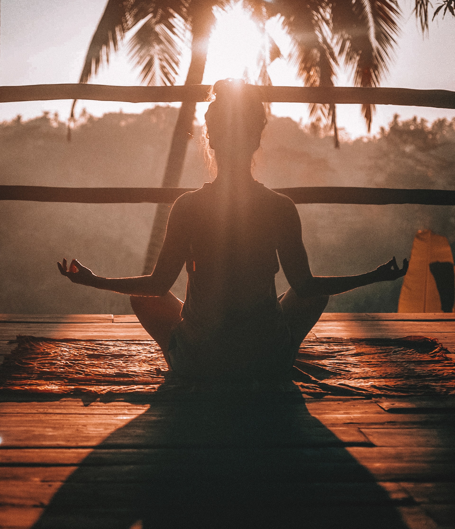 A person sits cross-legged on a mat in a meditative pose, facing sunrise with palm trees in the background. Sunlight streams through, generating health and creating a peaceful, serene atmosphere.
