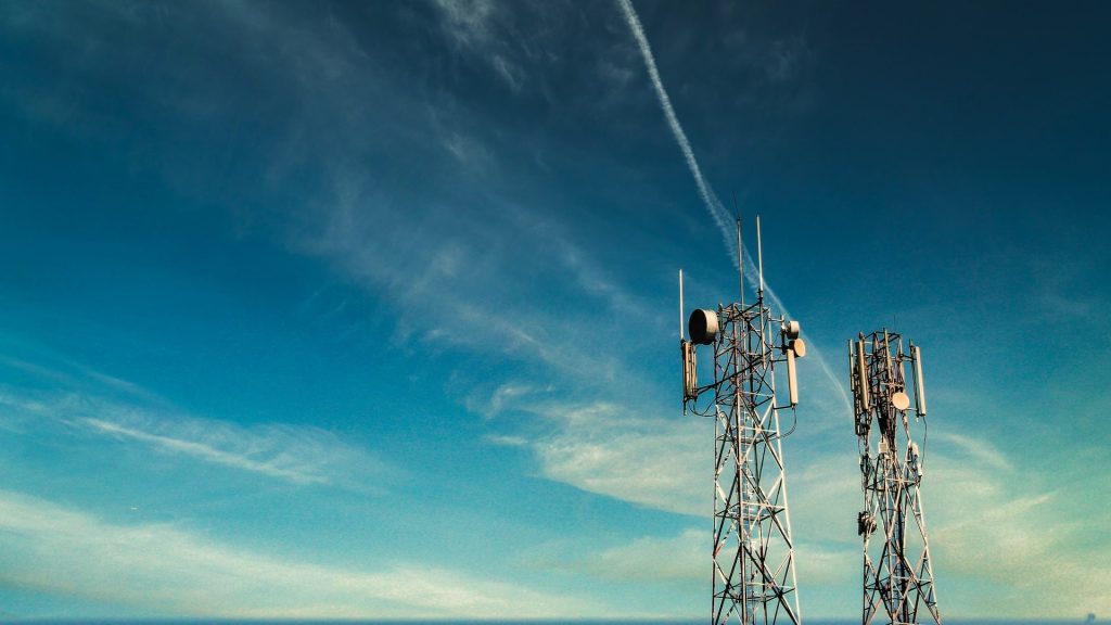 Two tall cell towers with antennas stand against a blue sky with wispy clouds and a visible jet contrail, highlighting the presence of modern 5G telecommunication technology.