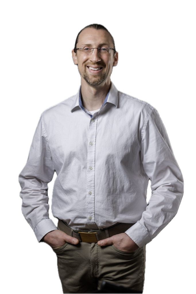 Brandon LaGreca smiling, wearing wire-rimmed glasses and a light grey button-down shirt, hands in pockets, white background.