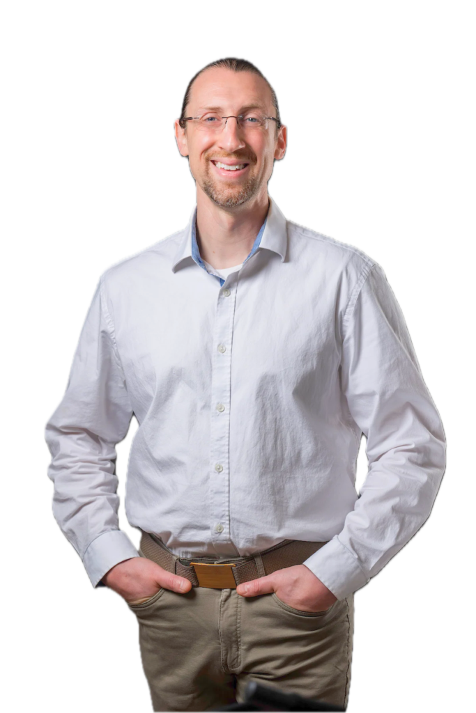 Brandon LaGreca smiling, wearing wire-rimmed glasses and a light grey button-down shirt, hands in pockets, white background.